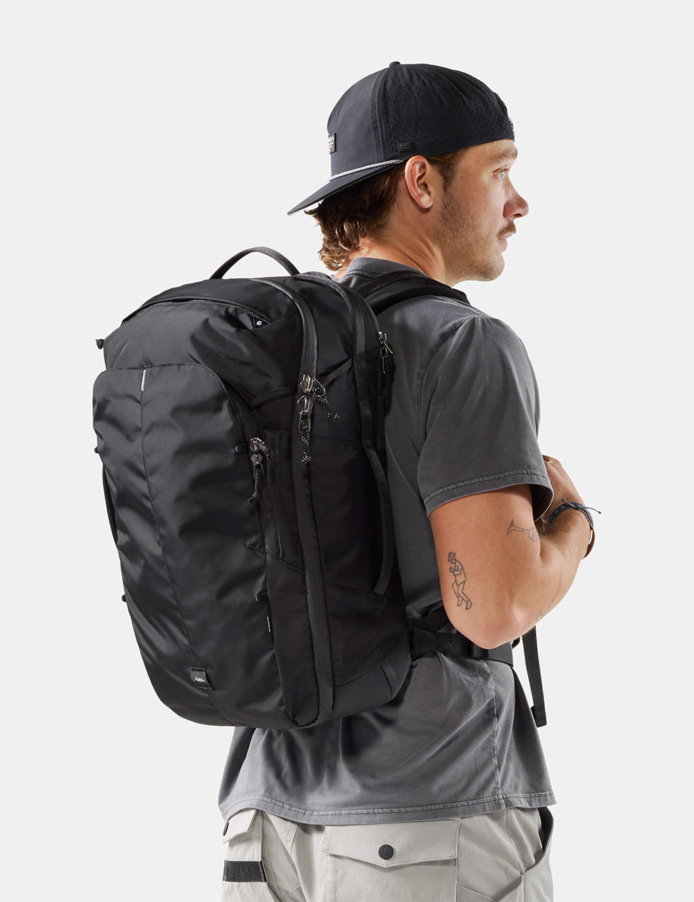 Matador 30L Carry-On Travel Pack in Black - a man wearing a black baseball cap, gray t-shirt, and light gray cargo pants is shown from a side-back angle, wearing a black technical backpack. The photo is taken against a clean, light gray studio background