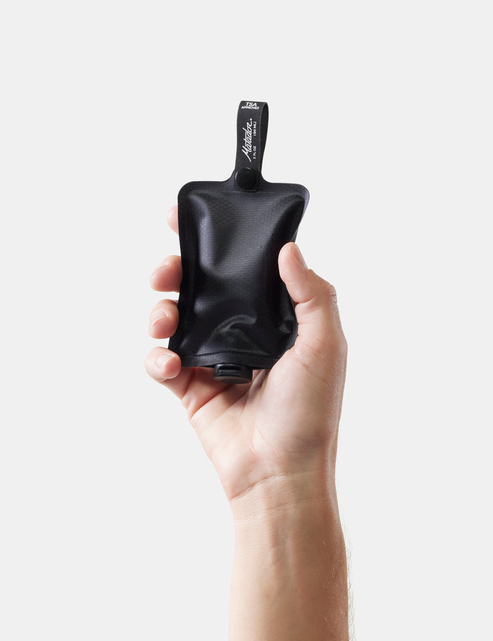 Matador FlatPak™ Toiletry Bottles - 3 Pack (Multi-Colour) - hand holding a black toiletry bottle on a white background