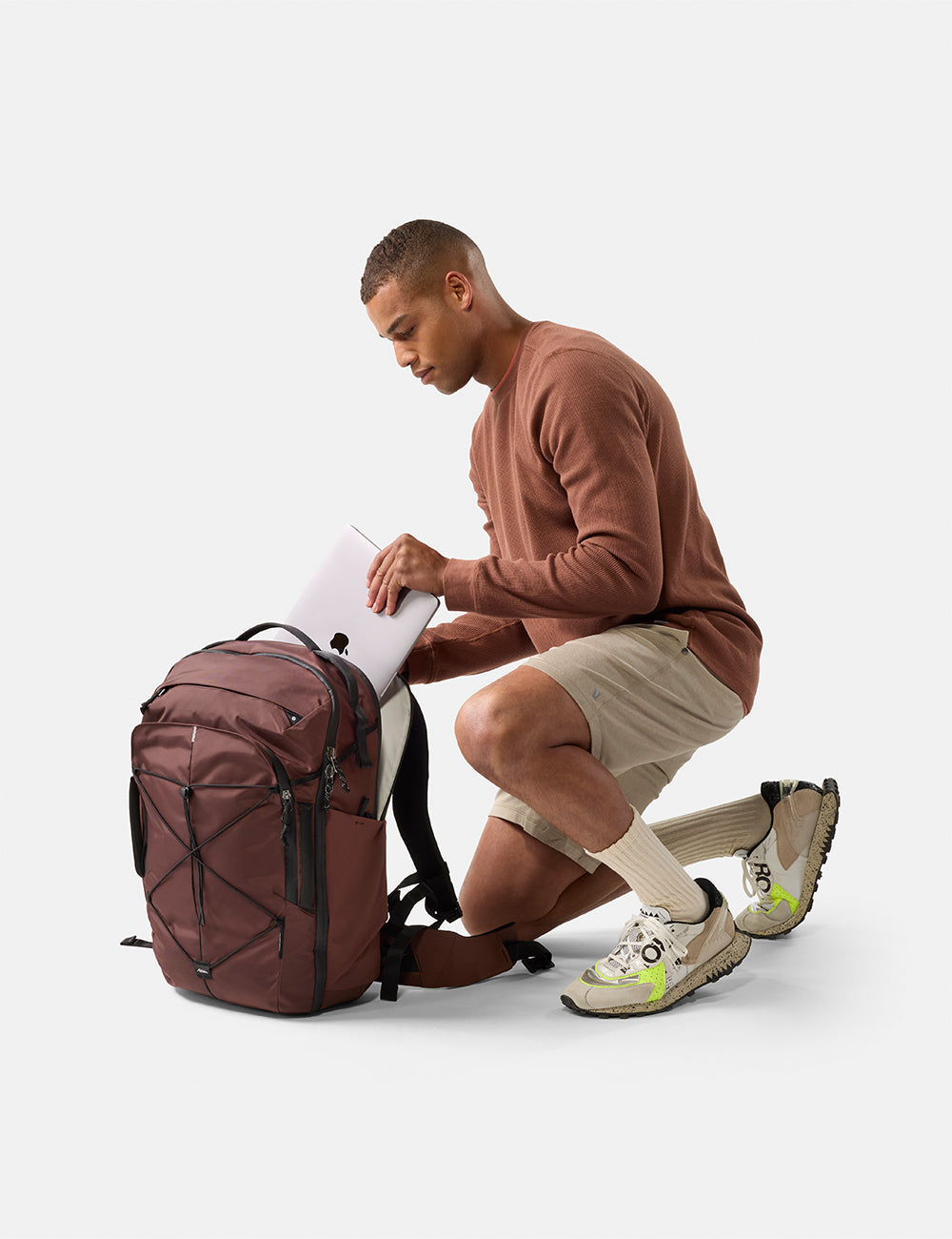 Matador 35L Global Travel Pack in Garnet - man putting laptop in garnet backpack, wearing a brown sweater and beige shorts, on a white background