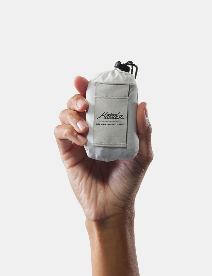 Matador Mini Pocket Blanket™ 4.0 - 110 x 70cm in Ghost Grey - hand holding a small white bag with a brand logo on a light gray background