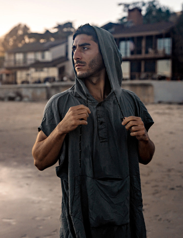 Matador NanoDry™ Packable Towel Poncho in Charcoal - man wearing a hooded garment on a beach with buildings in the background